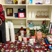 A festive hot chocolate bar setup on a table with a holiday-themed cloth. A sign reads "Hot Chocolate Bar" atop a dispenser with a large red and black checkered bow. Surrounding items include stacks of white cups, two bottles of Monin syrup, a small Christmas tree, and various toppings like marshmallows, chocolate chips, and candy canes, each labeled with a tent card. A framed sign on the right encourages warming up with a cup of hot chocolate.