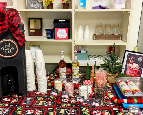 A festive hot chocolate bar setup on a table with a holiday-themed cloth. A sign reads "Hot Chocolate Bar" atop a dispenser with a large red and black checkered bow. Surrounding items include stacks of white cups, two bottles of Monin syrup, a small Christmas tree, and various toppings like marshmallows, chocolate chips, and candy canes, each labeled with a tent card. A framed sign on the right encourages warming up with a cup of hot chocolate.