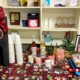 A festive hot chocolate bar setup on a table with a holiday-themed cloth. A sign reads "Hot Chocolate Bar" atop a dispenser with a large red and black checkered bow. Surrounding items include stacks of white cups, two bottles of Monin syrup, a small Christmas tree, and various toppings like marshmallows, chocolate chips, and candy canes, each labeled with a tent card. A framed sign on the right encourages warming up with a cup of hot chocolate.