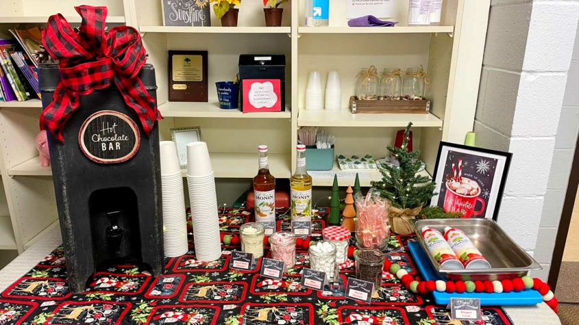 A festive hot chocolate bar setup on a table with a holiday-themed cloth. A sign reads "Hot Chocolate Bar" atop a dispenser with a large red and black checkered bow. Surrounding items include stacks of white cups, two bottles of Monin syrup, a small Christmas tree, and various toppings like marshmallows, chocolate chips, and candy canes, each labeled with a tent card. A framed sign on the right encourages warming up with a cup of hot chocolate.