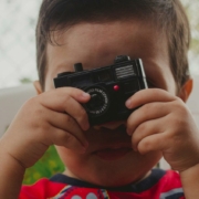 A young child is holding a small toy camera up to their face, pretending to take a photo. The camera lens is centered over the child's right eye, and the top of the child's head and background are softly out of focus, creating a candid and playful atmosphere.