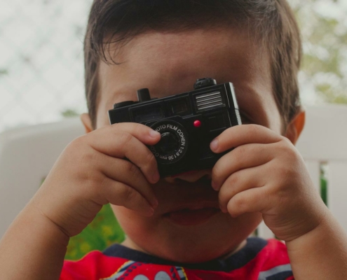A young child is holding a small toy camera up to their face, pretending to take a photo. The camera lens is centered over the child's right eye, and the top of the child's head and background are softly out of focus, creating a candid and playful atmosphere.