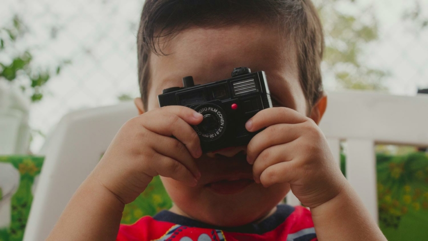 A young child is holding a small toy camera up to their face, pretending to take a photo. The camera lens is centered over the child's right eye, and the top of the child's head and background are softly out of focus, creating a candid and playful atmosphere.