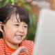 A young girl with black hair in pigtails is smiling while looking at a laptop screen. She is wearing a red and white striped shirt and has black earbuds in her ears. The background is softly blurred with greenery, suggesting she is outdoors.