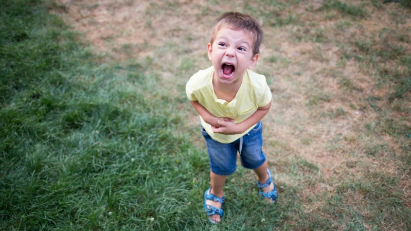 A young boy in a yellow shirt and blue shorts stands on a patchy grassy lawn, shouting with an exaggerated facial expression. He holds his stomach as if pretending to be in distress or acting playful. His sandals are blue, and his posture is slightly leaned forward, emphasizing his animated demeanor.